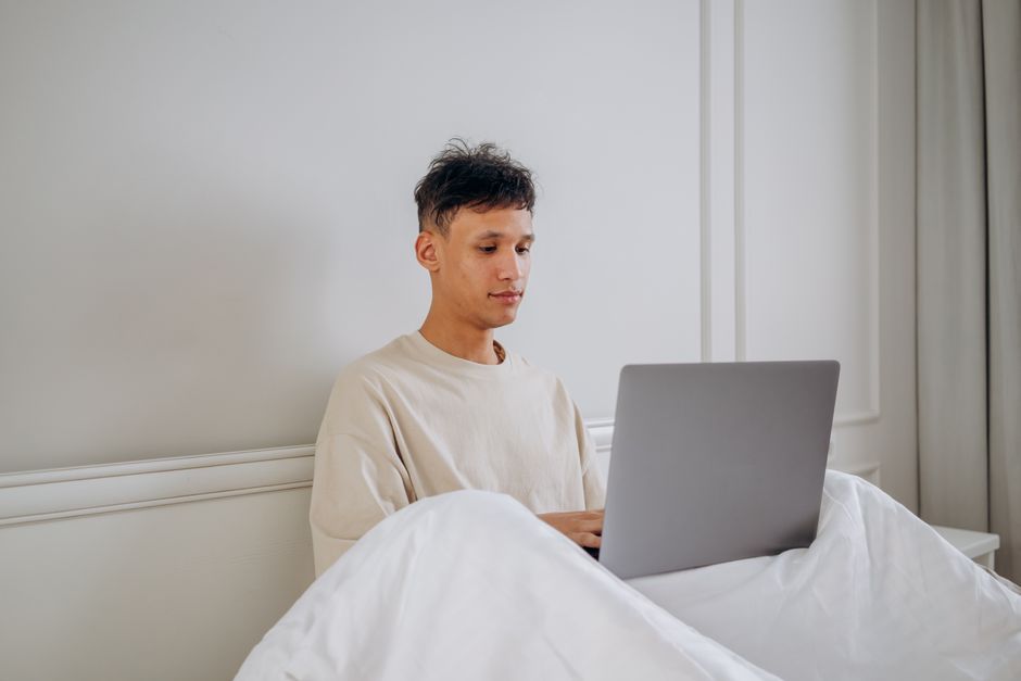 Young man in bed using laptop, representing remote work and weekday business travel